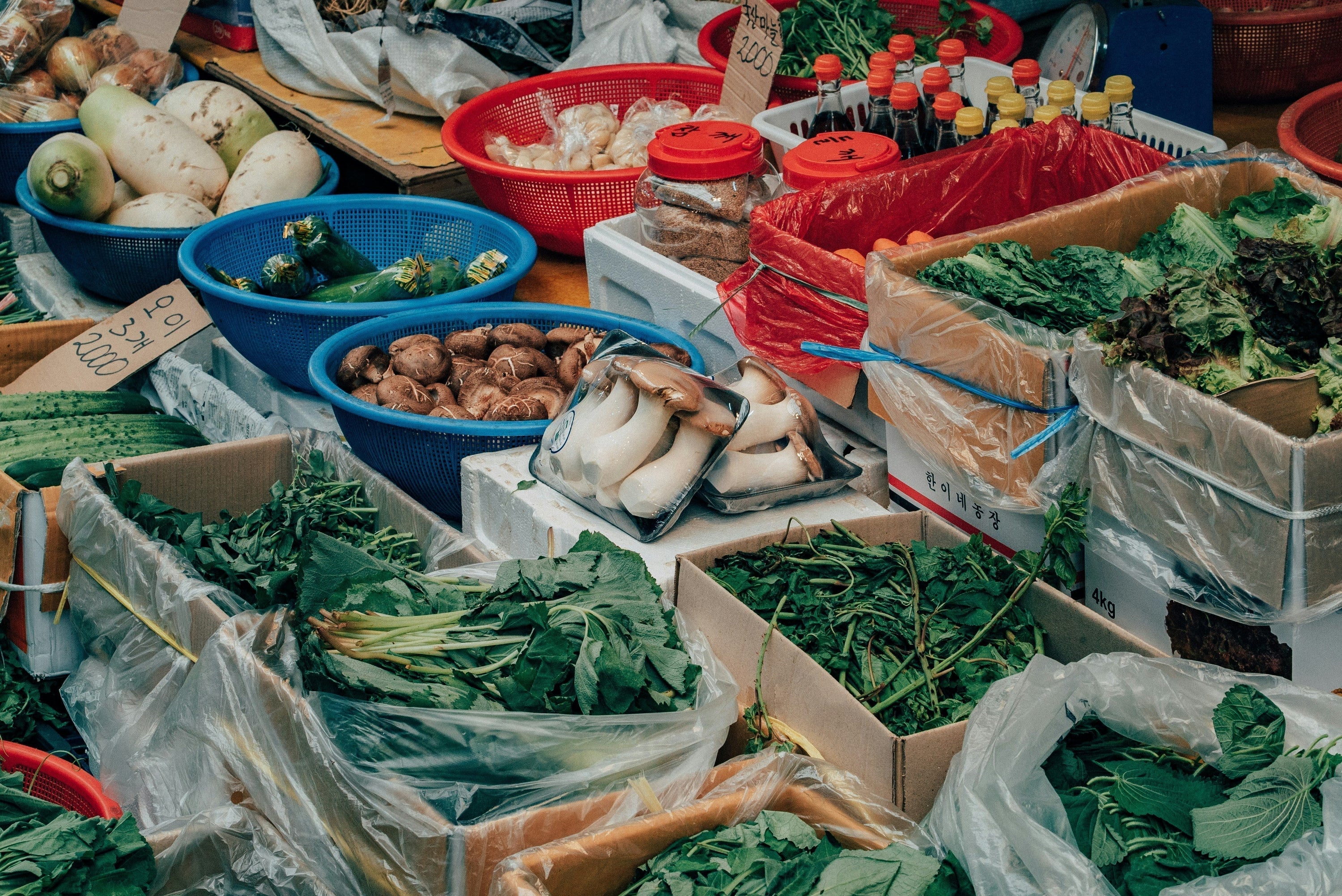 Fresh greens, mushrooms, roots, and bottled condiments displayed in crates and baskets at an outdoor market in Korea.