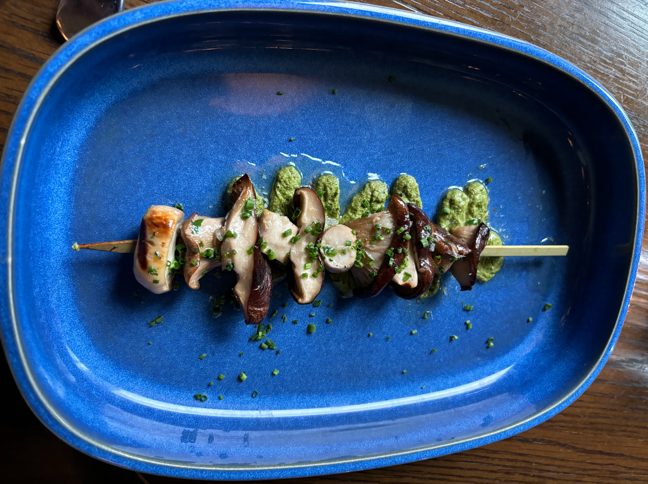 Grilled mushroom skewer with chives over a green herb purée, served on a bright blue plate.