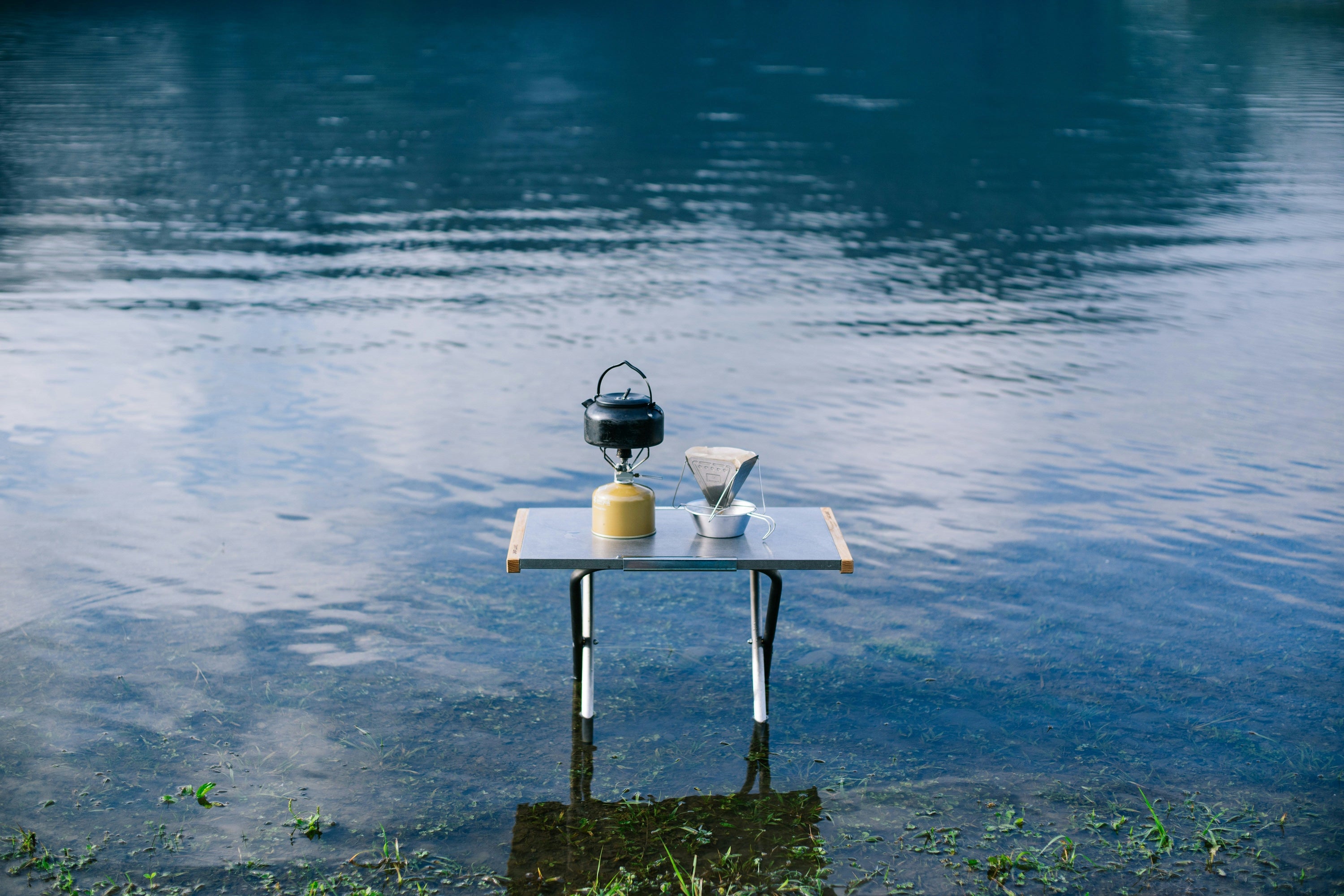 Small table standing in shallow lake water with a kettle and pour-over coffee setup
